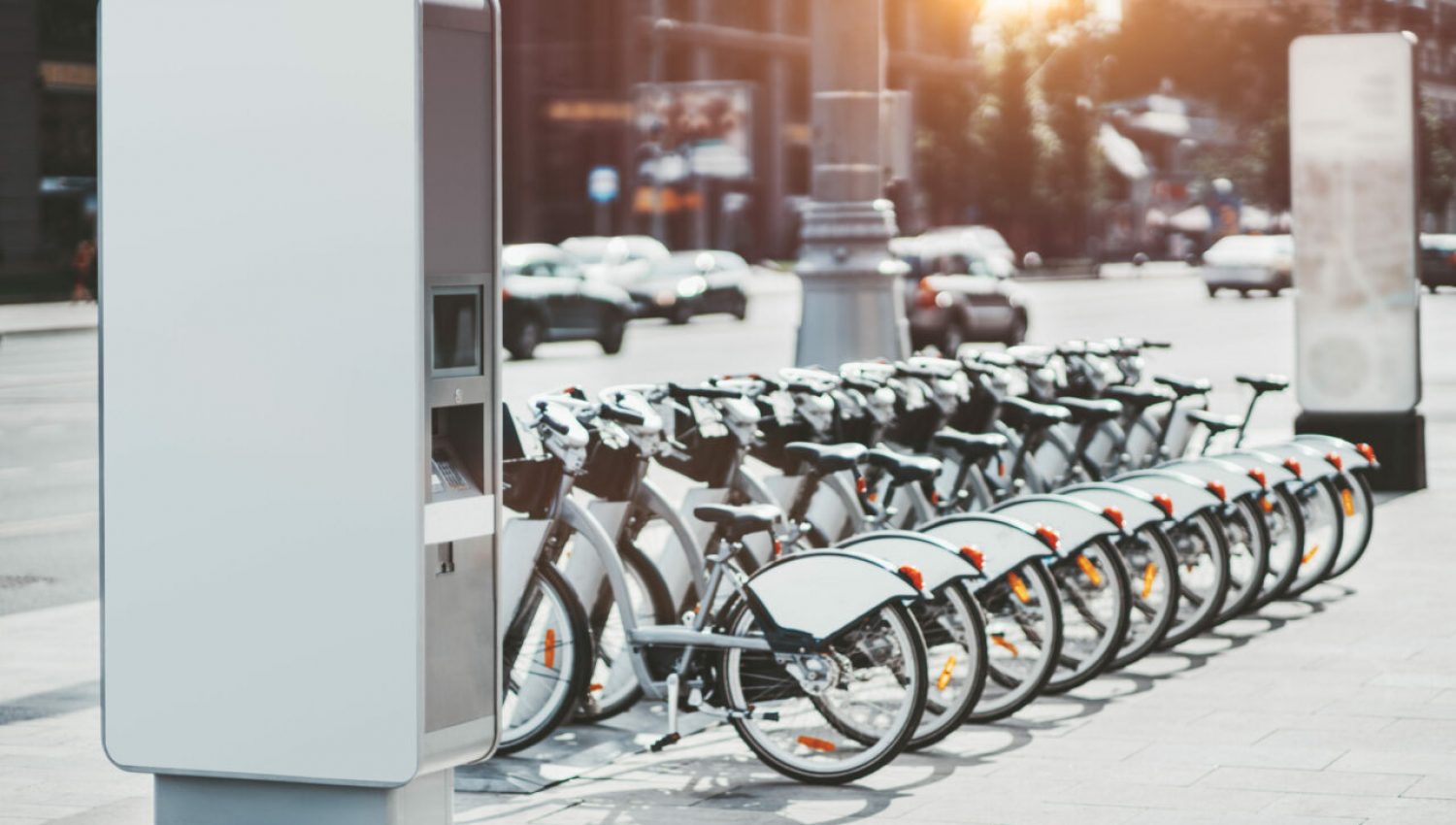 Modern rental electric bikes standing a row during charging on a bright day with a selective focus on the vertical payment terminal with a mock-up of informational or an ad banner placeholder on it